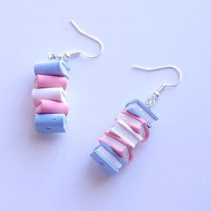 May include: A pair of dangle earrings featuring a stack of miniature books in shades of blue, pink, and white. The books are made of polymer clay and are attached to silver-toned earring hooks.