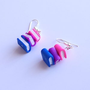 May include: A pair of earrings featuring a stack of three miniature books. The books are painted in shades of blue, purple, and pink. The earrings are made with silver hooks.