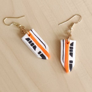 May include: A pair of dangle earrings featuring a white, orange, and black design of a monorail train. The earrings have gold hooks.