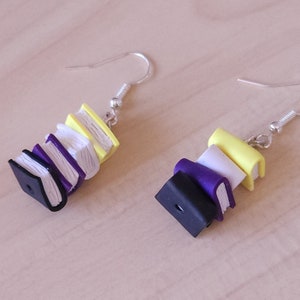 May include: A pair of dangle earrings featuring a stack of miniature books in black, purple, white, and yellow. The books are stacked on top of each other and have a realistic texture.