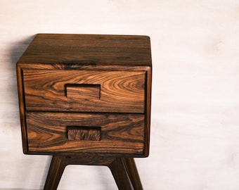 May include: A dark brown wooden nightstand with two drawers and a mid-century modern design. The nightstand has a rectangular top and a base with angled legs. The wood grain is visible throughout the piece.