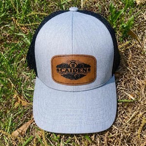 May include: A light gray trucker hat with a black mesh back and a curved brim. The hat features a brown leather patch with the text "$CAIDEN$" and a crown design.
