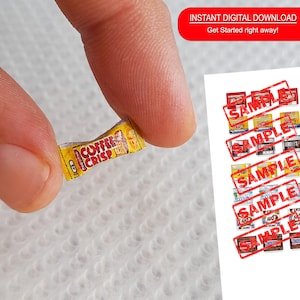 Puede incluir: Una réplica en miniatura de un envoltorio de chocolate Coffee Crisp. El envoltorio es amarillo con texto rojo y blanco. El envoltorio está siendo sostenido por un dedo.