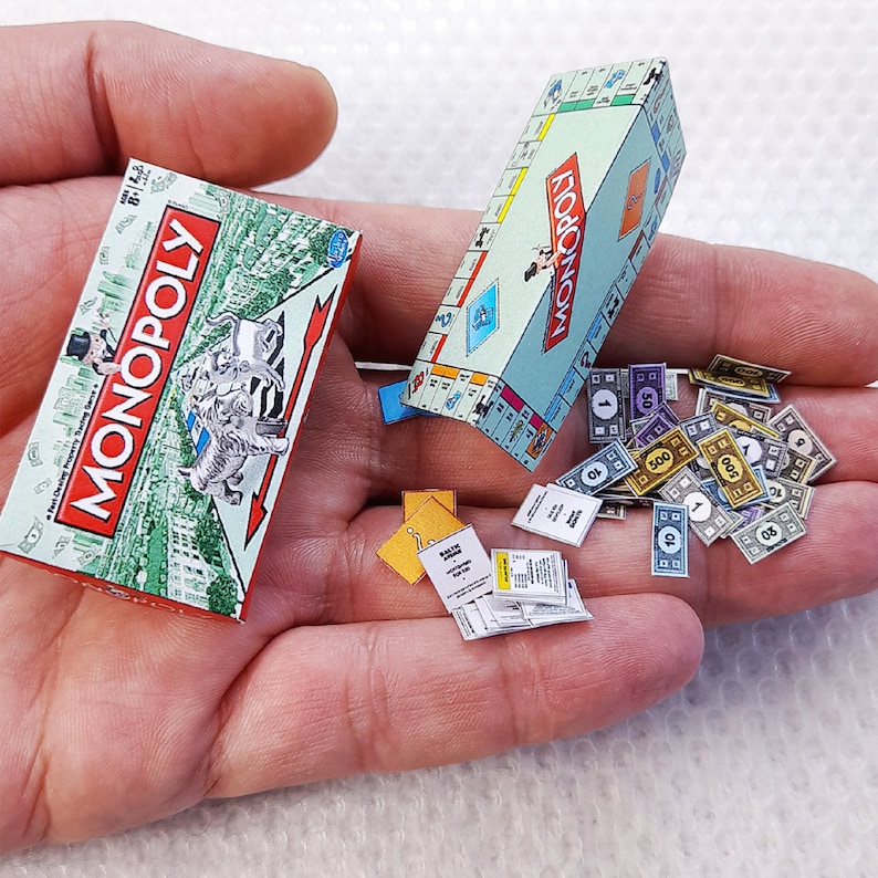 May include: Miniature Monopoly game set, including a game board, box, play money, and chance cards. The box features the Monopoly logo in red and white, with a cityscape background. The game board is light blue with colorful property spaces.