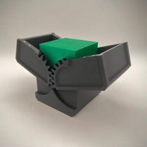 May include: A black plastic card storage box with a green deck of cards inside. The box has a gear mechanism on the side.
