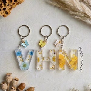 May include: Resin keyrings spelling "VHN". Each letter is filled with dried flowers and gold flakes. The keyrings have silver rings and are decorated with small butterfly charms in blue, yellow, and white.
