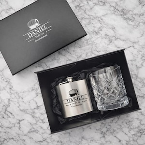 Könnte beinhalten: Eine schwarze Geschenkbox mit einem silbernen Metallflachmann und einem klaren Kristallglas. Der Flachmann und der Deckel der Box sind mit einem Fass-Symbol, dem Namen DANIEL, dem Datum 26.07.26 und dem Wort Groomsman graviert.