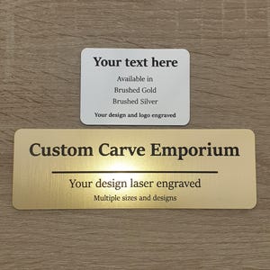 May include: A gold metal sign with the text "Custom Carve Emporium" and the phrase "Your design laser engraved" in black lettering. A smaller white sign above reads "Your text here" with the text "Available in Brushed Gold Brushed Silver Your design and logo engraved".