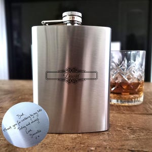 May include: A silver-coloured hip flask with the name "David" engraved in a decorative frame. A small, round metal piece with handwritten text is in the foreground. A glass of amber liquid sits nearby.