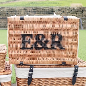 May include: A rectangular light brown wicker picnic basket featuring the black letters E&R centred on the front. The basket has a hinged lid with black leather straps and a white fabric lining visible along the top edge and the base section.