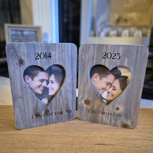 May include: A grey wooden photo frame with two hinged panels, each with a heart-shaped aperture. The left panel reads "2014" and "Our First Date", the right panel reads "2025" and "Our Forever Date". The frame displays photos of a couple.