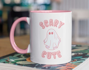 Puede incluir: Taza de cerámica blanca con borde y asa de color rosa. La taza presenta un fantasma rosa con el texto "Scary Cute" en letras rosas.