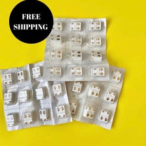 Puede incluir: Primer plano de varios paquetes de pequeños pendientes magnéticos redondos, blancos con detalles dorados. Los paquetes están apilados sobre una superficie amarilla.