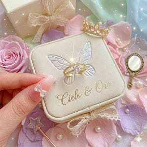 May include: A gold ring with a square, clear stone sits on a cream-colored ring box with a butterfly design. The box has the text "Cielo & Oro". A small gold crown and a hand mirror are also visible.