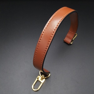 May include: A brown leather bag handle with gold-colored hardware. The handle is curved and stitched with a contrasting thread. The hardware includes a swivel clasp and a rectangular connector.