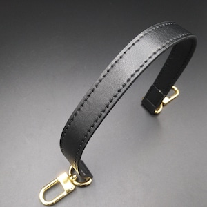 May include: A black leather handbag handle with gold-colored metal clasps. The handle is stitched along the edges and has a curved design. The clasps are in a U-shape.
