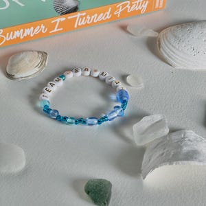 May include: A beaded bracelet with the text "TEAM JEREMIAH" in white letter beads, interspersed with blue beads. The bracelet is surrounded by seashells, sea glass, and a book titled "Summer I Turned Pretty".