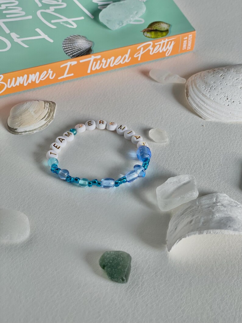 May include: A beaded bracelet with the text "TEAM JEREMIAH" in white letter beads, interspersed with blue beads. The bracelet is surrounded by seashells, sea glass, and a book titled "Summer I Turned Pretty".