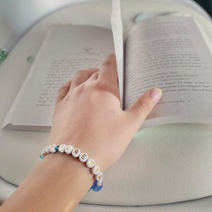 May include: A beaded bracelet spelling "JEREMIA" in white letter beads is worn on a wrist, next to an open book. The bracelet has blue and white accent beads. The book's pages are open, with the hand turning a page.