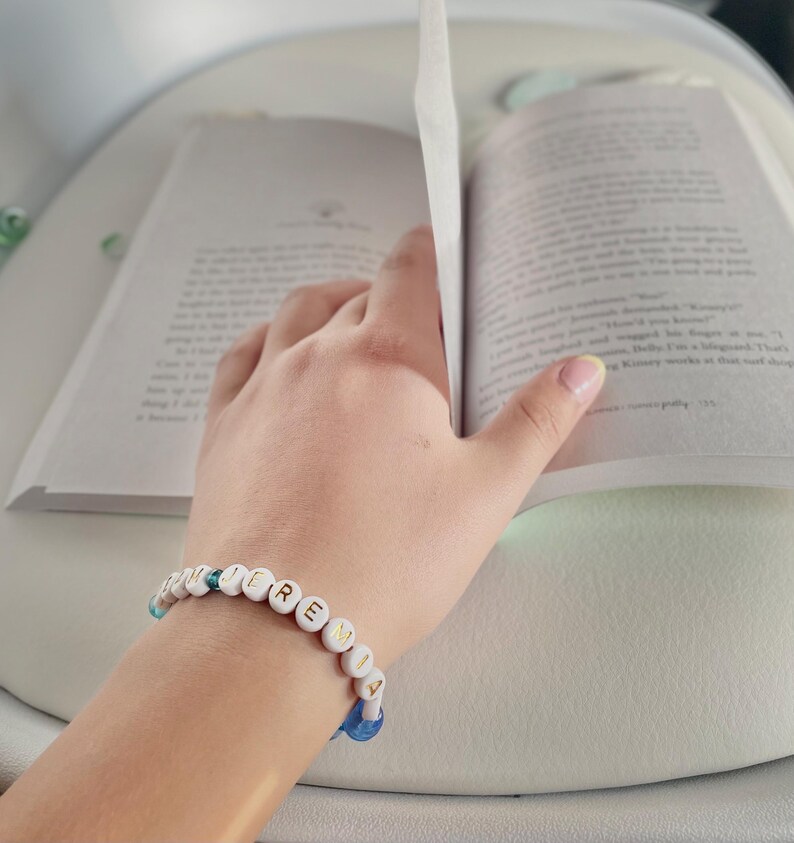 May include: A beaded bracelet spelling "JEREMIA" in white letter beads is worn on a wrist, next to an open book. The bracelet has blue and white accent beads. The book's pages are open, with the hand turning a page.