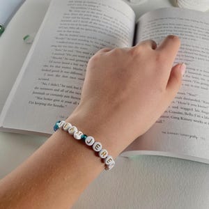 May include: A beaded bracelet spelling out "TEAM JERE" in white letter beads. The bracelet is on a wrist, with a book open in the background. The bracelet has blue and green beads.