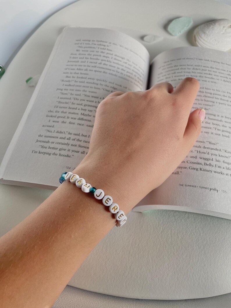 May include: A beaded bracelet spelling out "TEAM JERE" in white letter beads. The bracelet is on a wrist, with a book open in the background. The bracelet has blue and green beads.