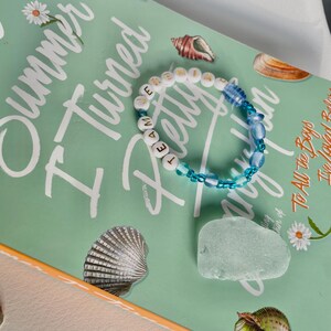 May include: A beaded bracelet with the word "TEAM" spelled out in white letter beads. The bracelet also features blue beads and is set against a book cover with the title "I Turned Pretty". Seashells and a piece of sea glass are also visible.