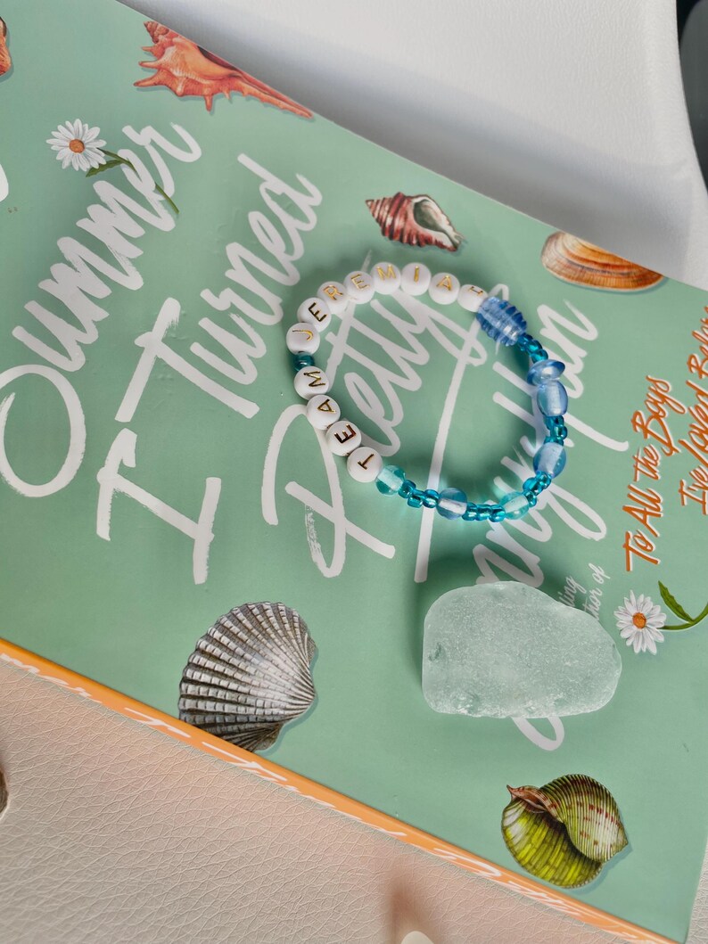 May include: A beaded bracelet with the word "TEAM" spelled out in white letter beads. The bracelet also features blue beads and is set against a book cover with the title "I Turned Pretty". Seashells and a piece of sea glass are also visible.