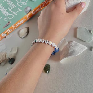 May include: A white beaded bracelet spelling out "Jeremiah" with white letter beads and blue accent beads. The bracelet is on a wrist, next to a book titled "The Summer I Turned Pretty" and seashells.