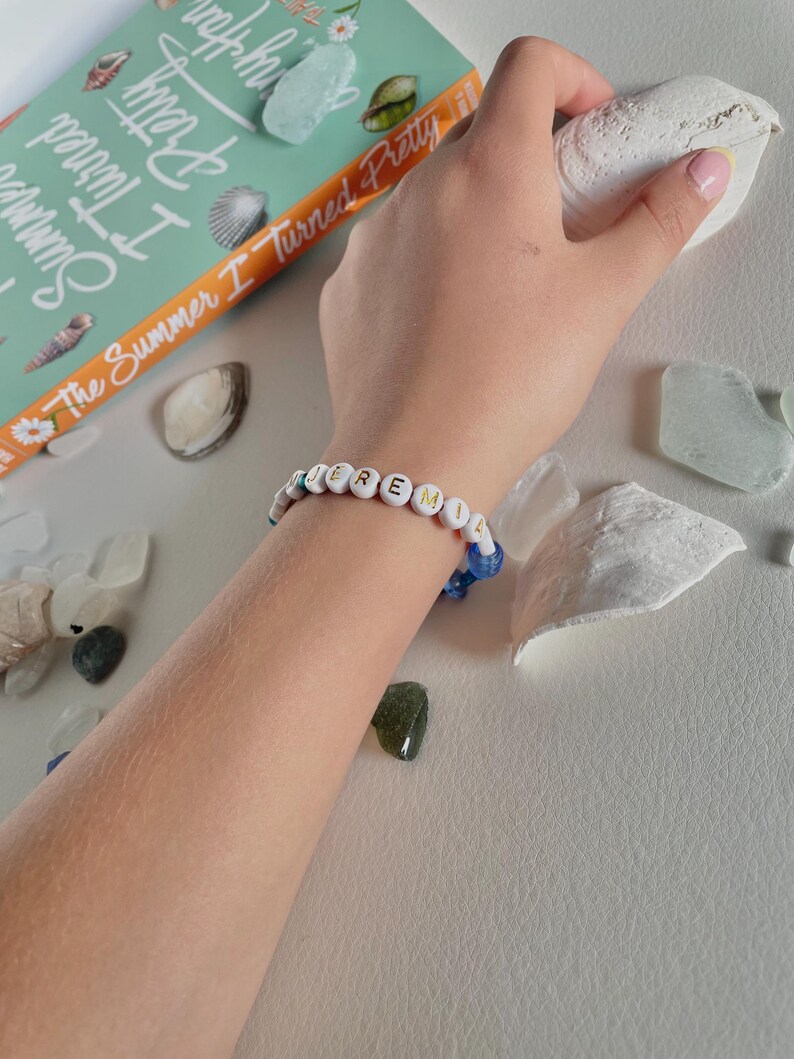 May include: A white beaded bracelet spelling out "Jeremiah" with white letter beads and blue accent beads. The bracelet is on a wrist, next to a book titled "The Summer I Turned Pretty" and seashells.