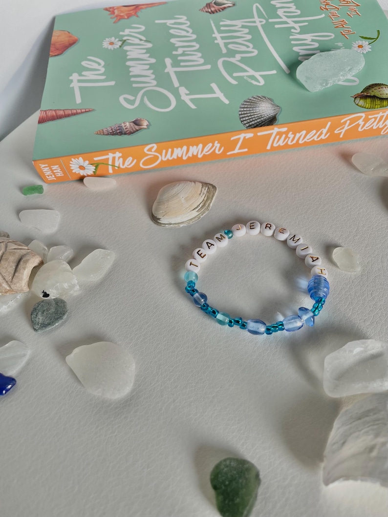 May include: A beaded bracelet with the word "TEAM JEREMIAH" spelled out in white and blue beads. The bracelet is surrounded by sea glass, seashells, and a book titled "The Summer I Turned Pretty."