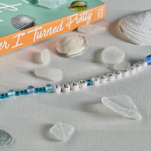 May include: A still life featuring a book titled "Summer I Turned Pretty" with seashells and sea glass. A beaded bracelet spells out "TEAM JEREMIA". The book has an orange spine and a teal cover. The sea glass is translucent and the shells are white.