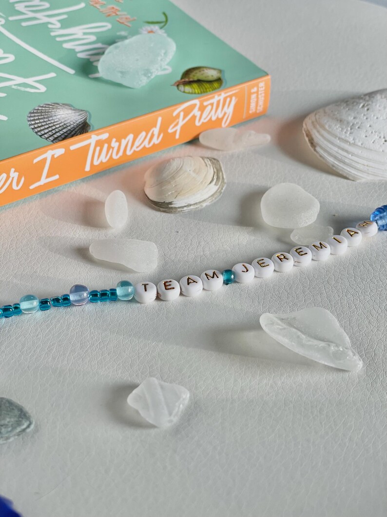 May include: A still life featuring a book titled "Summer I Turned Pretty" with seashells and sea glass. A beaded bracelet spells out "TEAM JEREMIA". The book has an orange spine and a teal cover. The sea glass is translucent and the shells are white.