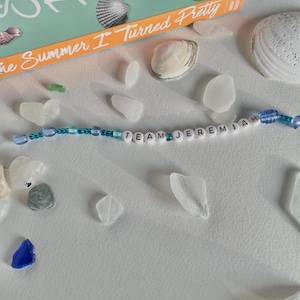 May include: A beaded bracelet with the text "TEAM JEREMIAH" is displayed with seashells, sea glass, and a book titled "The Summer I Turned Pretty". The bracelet features white letter beads and blue beads.