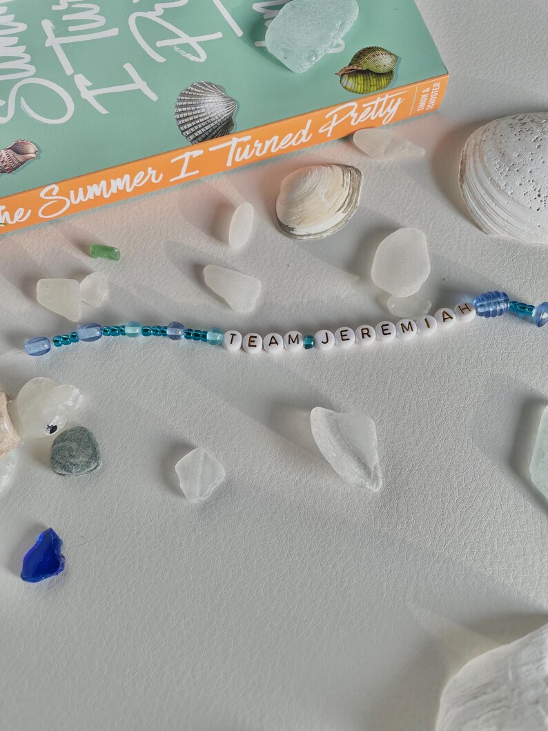 May include: A beaded bracelet with the text "TEAM JEREMIAH" is displayed with seashells, sea glass, and a book titled "The Summer I Turned Pretty". The bracelet features white letter beads and blue beads.