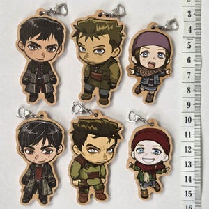 Op de afbeelding: Zes houten sleutelhangers met anime-stijl personages en metalen sluitingen. De personages omvatten twee mannen in donkere jassen, twee mannen in olijfgroene outfits en twee figuren met wintermutsen. Een verticaal meetlint toont een hoogte van 15 cm.