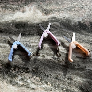 May include: Three small, colorful, keychain scissors. The scissors are pink, blue, and beige. Each scissor has a silver blade and a small silver chain.