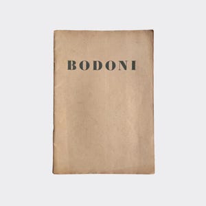May include: A rectangular book with a weathered, light brown paper cover. The word BODONI is printed in a bold, dark gray, serif typeface across the upper center of the front cover. The paper texture appears aged with subtle variations in color.
