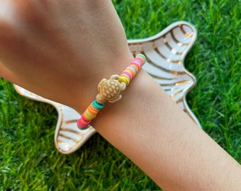 Könnte beinhalten: Ein farbenfrohes Perlenarmband mit einem kleinen Schildkrötenanhänger. Das Armband besteht aus kleinen, runden Perlen in verschiedenen Farben, darunter Pink, Gelb, Grün, Blau und Orange. Der Schildkrötenanhänger ist aus weißem Kunststoff und mit einem Metallring am Armband befestigt.