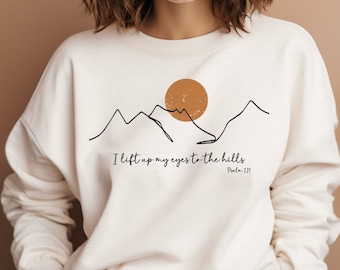 May include: White sweatshirt with a black line drawing of mountains and a sun. The text "I lift up my eyes to the hills Psalm 121" is printed below the image.