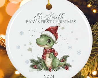 Könnte beinhalten: Ein weißer, runder Weihnachtsschmuck mit einer goldenen Aufhängeschlaufe. Er zeigt einen grünen Dinosaurier mit Weihnachtsmütze und Schal, mit dem Text "Eli Smith BABY'S FIRST CHRISTMAS 2024". Schneeflocken und Beeren sind ebenfalls abgebildet.