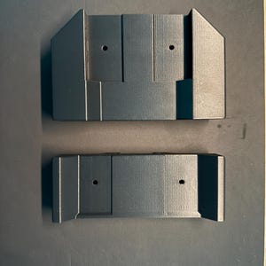 May include: Two black plastic wall mounts with two holes each. The mounts are designed to hold a device or object.