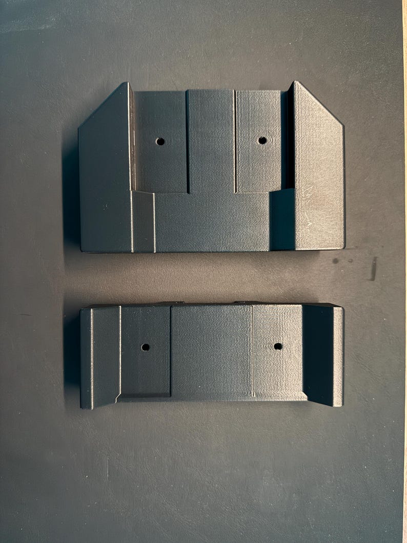 May include: Two black plastic wall mounts with two holes each. The mounts are designed to hold a device or object.