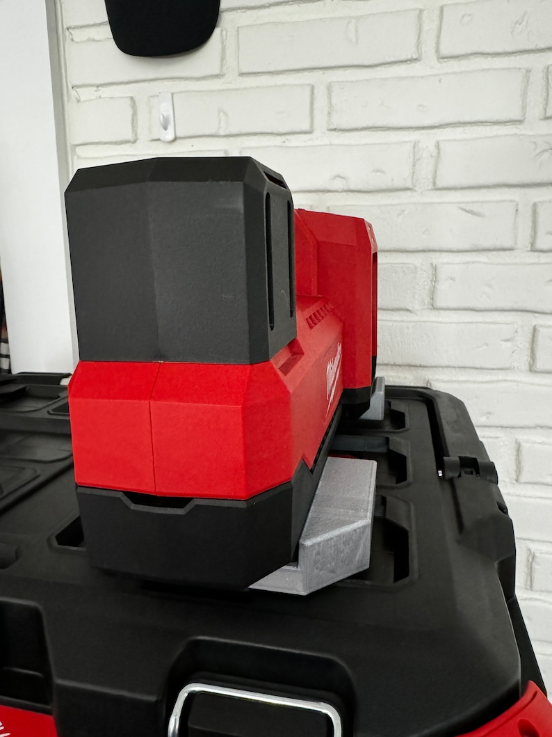 May include: A red and black power tool battery sits on top of a black tool box. The battery has a black handle and a red body with a black label that says "Milwaukee".