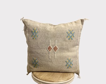 May include: A beige throw pillow with a geometric pattern in shades of blue, yellow, and red. The pillow is made of a soft, woven fabric and has a fringed edge.
