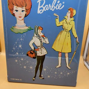May include: A light blue metal Barbie carrying case featuring a vintage illustration of a woman with red hair, a white pearl necklace, and two full-body figures in stylish outfits. The text Barbie is printed in white, with 1963 Mattel, Inc. at the bottom.