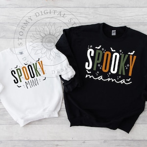 Pode incluir: Duas blusas de frio brancas e pretas com tema de Halloween. A blusa branca tem o texto "Spooky Mini" em laranja, verde e branco com morcegos e estrelas. A blusa preta tem o texto "Spooky Mama" em laranja, verde e branco com morcegos e estrelas.