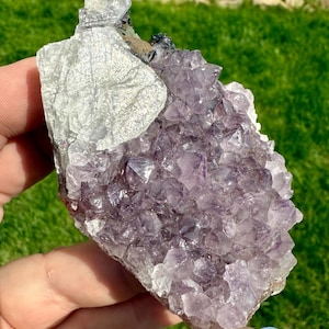 Puede incluir: Un grupo de cristales de amatista natural con una mezcla de cuarzo morado oscuro y claro. La formación de cristal tiene una superficie rugosa y texturizada. El cristal se sostiene sobre un fondo verde.