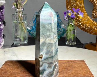 May include: A polished, hexagonal crystal tower featuring a blend of mossy green, grey, and white swirling patterns. The stone has a pointed top and a smooth, multi-faceted surface with visible natural inclusions and earthy, marbled color variations.