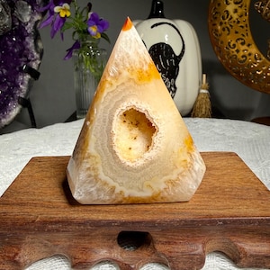 May include: A pyramid-shaped agate stone featuring concentric bands of cream, beige, and translucent white colors. The center of the stone contains a small, hollow geode cavity with a textured, crystalline interior and a bright orange tip.